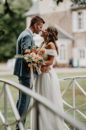 Un couple de mariés romantiques s'embrasse sur un balcon à balustrade blanche avec un magnifique bouquet floral pêche et orange.