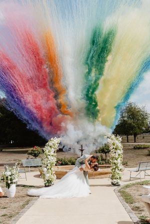 Bride and groom kiss under floral arch with rainbow colored powder smoke explosion at outdoor wedding ceremony.