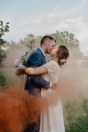 Un couple de mariés romantiques partage un baiser entouré d'effets de fumée colorés dans un jardin extérieur.