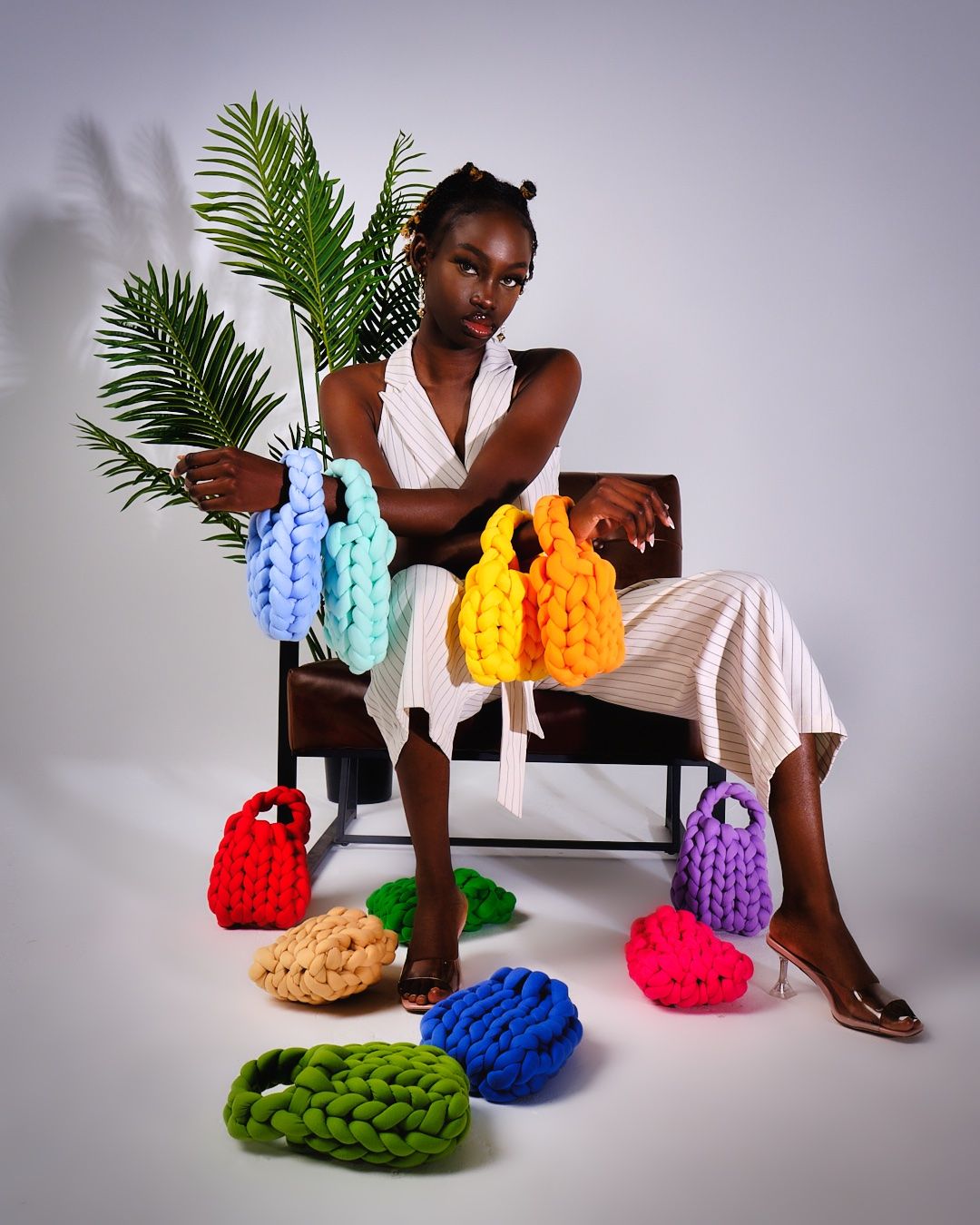 Colorful knitted handbags displayed with palm fronds against a grey background.