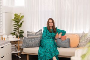 Someone in a green floral dress sits on a beige couch with decorative pillows and a fiddle leaf fig plant nearby.