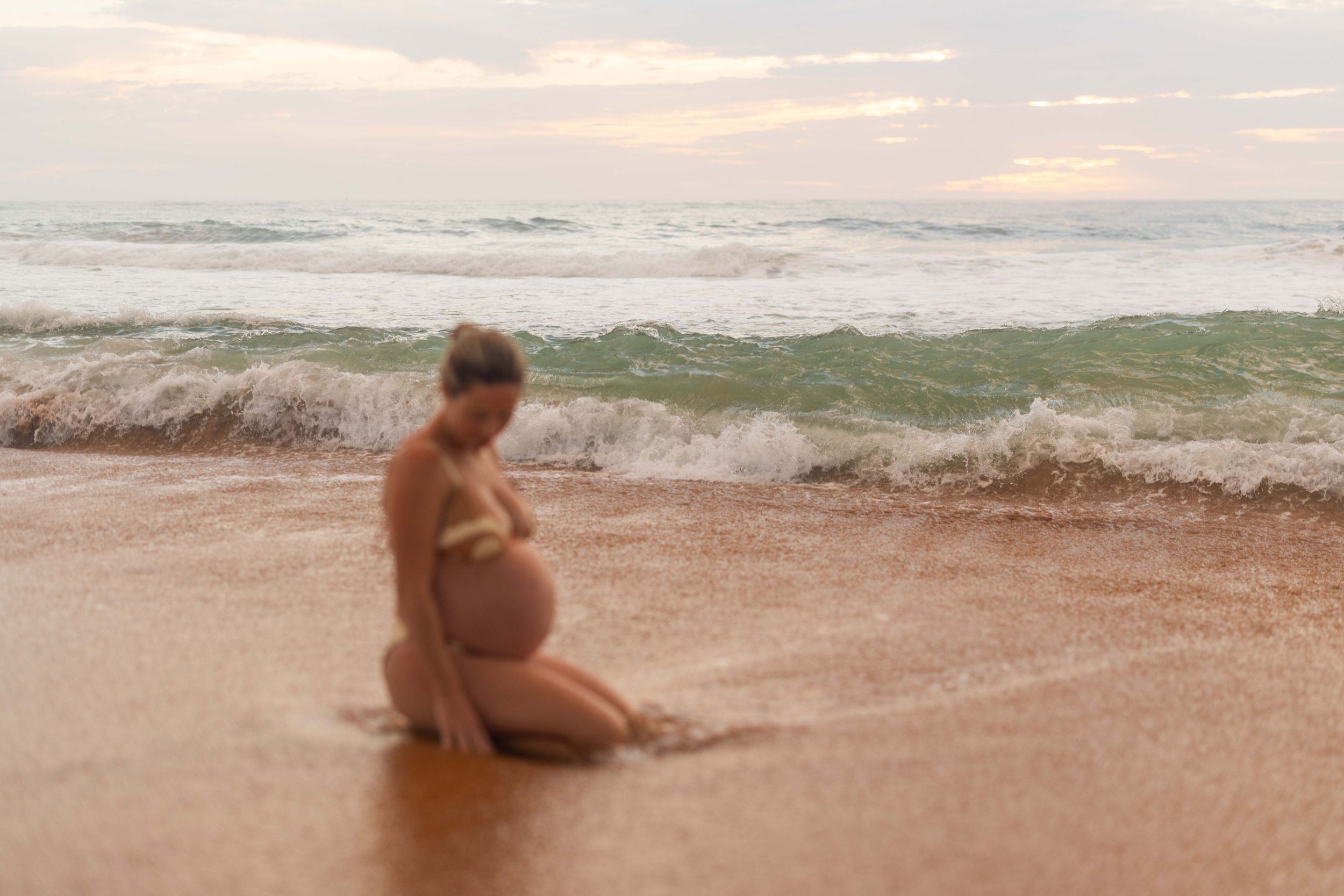 Pregnant woman in bikini sitting on sandy beach at sunset with ocean waves in the background.