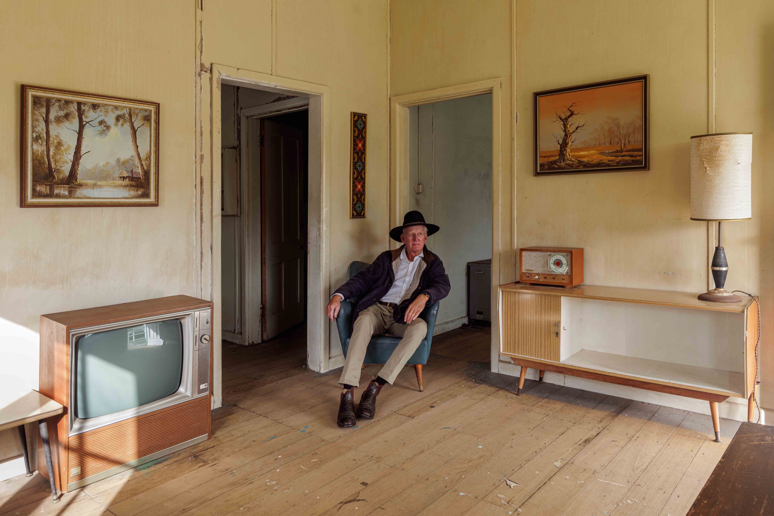 Man in cowboy hat sits in vintage room with retro TV, radio, and landscape paintings on worn wooden floors.