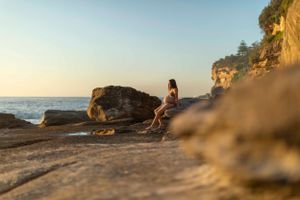 Person sitting on coastal rocks during golden hour with cliffs and ocean in background.