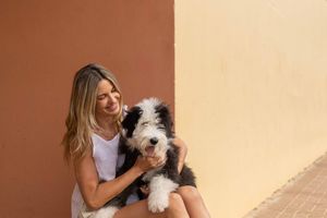 A person in a white top plays and cuddles with a small fluffy white dog against a beige background.