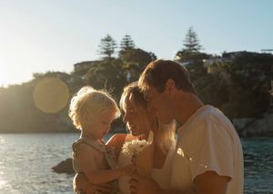 Family with young toddler sharing a tender moment by the water at golden hour with sunlit trees in background.