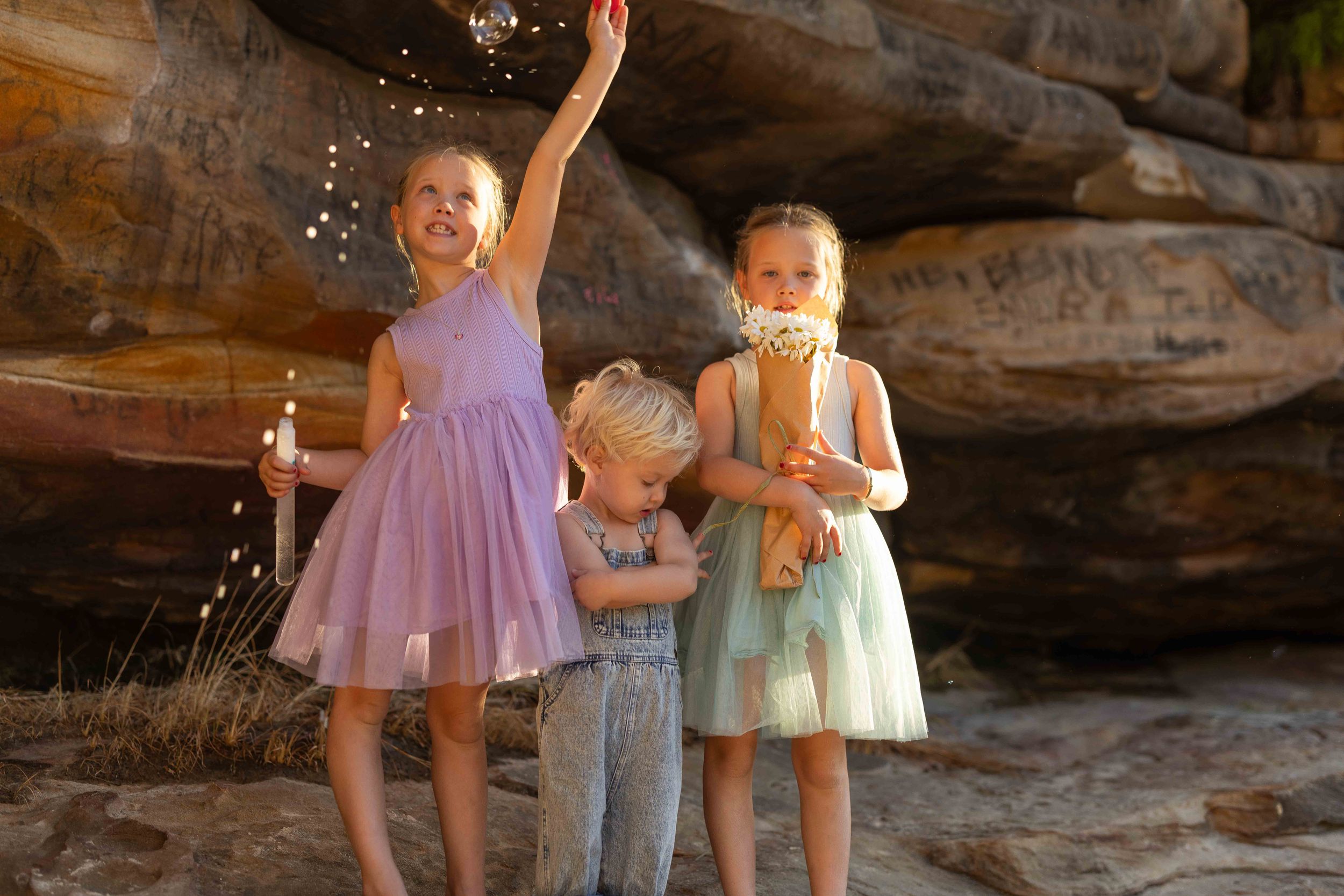 Three young children playing outdoors near sandstone rocks, with a girl in purple dress tossing confetti.