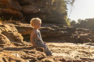 A young toddler with blonde hair sits on rocky sandy terrain outdoors, wearing denim overalls in warm golden sunlight.