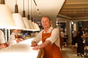 Chef in orange apron leaning on white restaurant counter under pendant lights in elegant dining room.