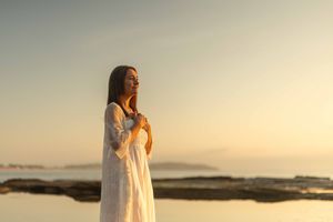 A person in a flowing white dress stands on a rocky beach at sunset creating a dreamy silhouette against the ocean.