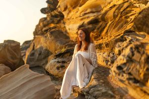 Woman in flowing white dress stands on rocky cliffs at sunset, creating a dreamy silhouette against golden light.