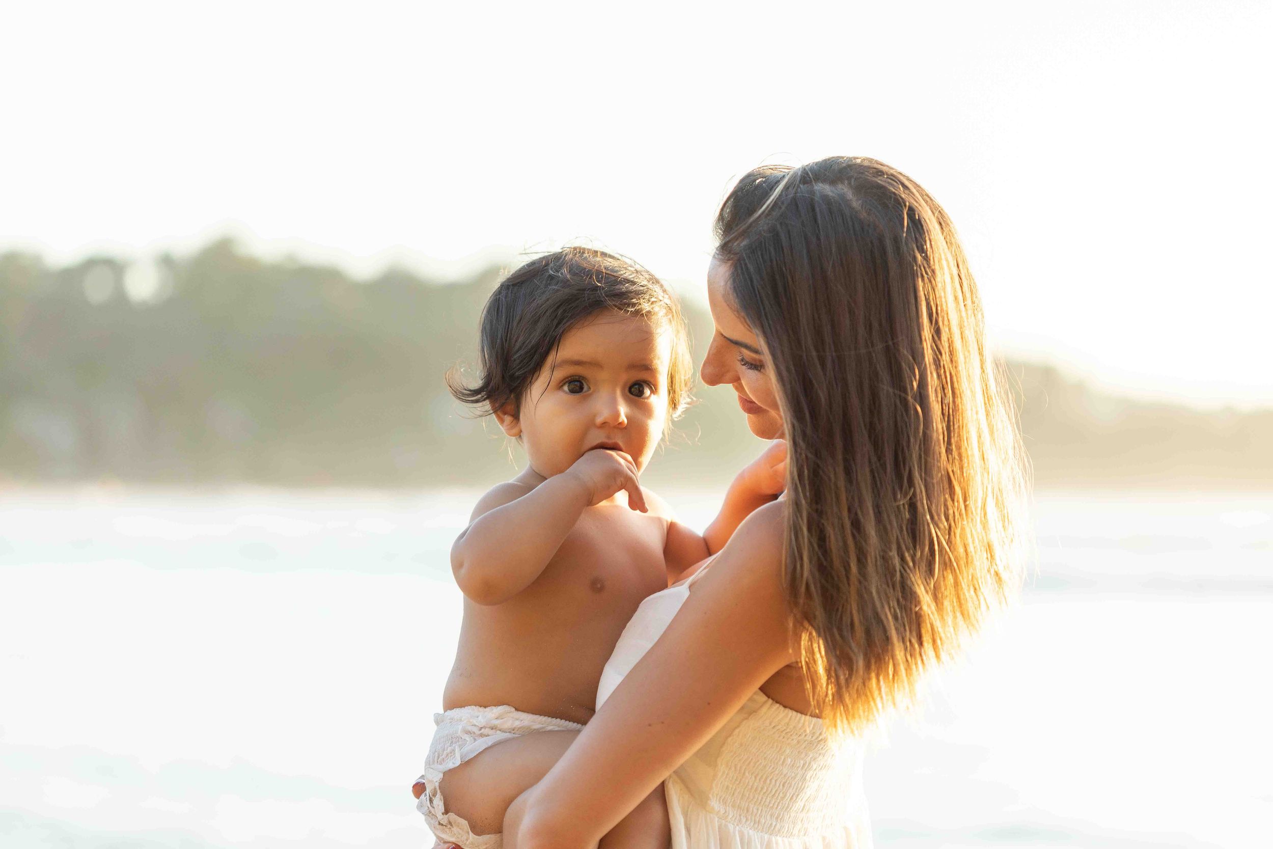 A mother tenderly holds her baby on a sunny beach, sharing a warm moment in golden light.