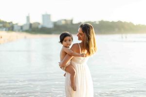 A mother in a white dress holds her toddler on a sunny beach with calm water and buildings in the background.