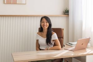 Professional working at modern wooden desk with laptop and coffee while smiling in bright home office setting.