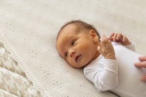 Adorable baby in white onesie lies on textured white blanket while reaching out with tiny hand.