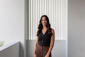 Professional wearing black sleeveless top and brown pants stands by window with vertical blinds in bright office space.