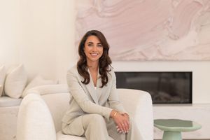 A woman in a beige blazer sits casually on a modern white sofa in a bright, minimalist setting.