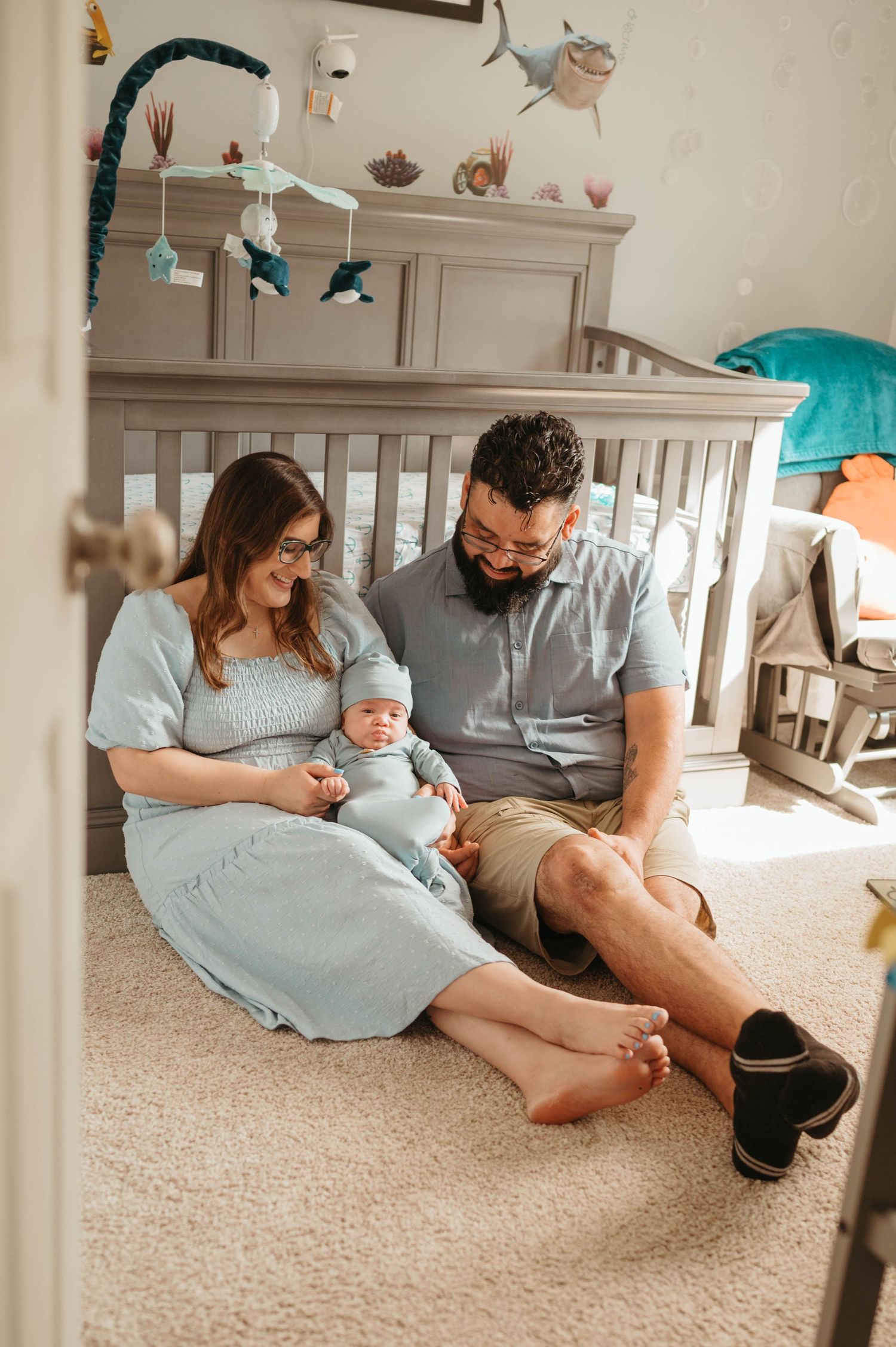 New parents in blue outfits sit on nursery carpet with newborn beside gray crib with ocean mobile.