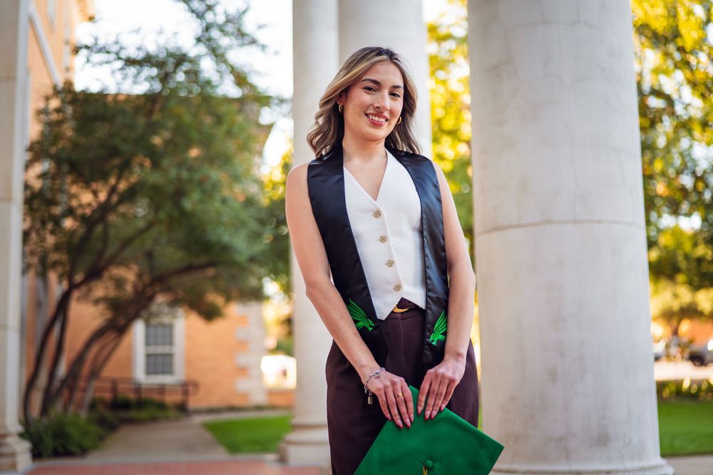Woman in black vest and green skirt poses elegantly by white stone columns during golden hour.