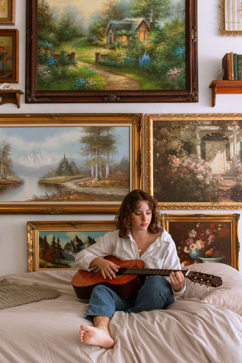 Girl sitting on a bed with a guitar surrounded by paintings hung on a wall