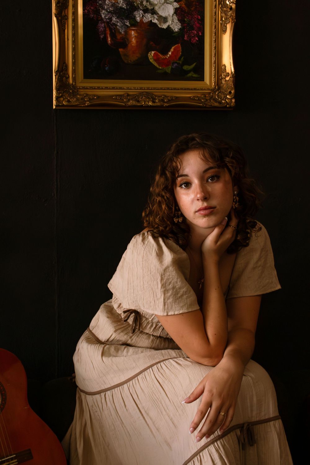 Girl looks into camera and sits on a couch with a guitar to the left of her and a gold frame above her