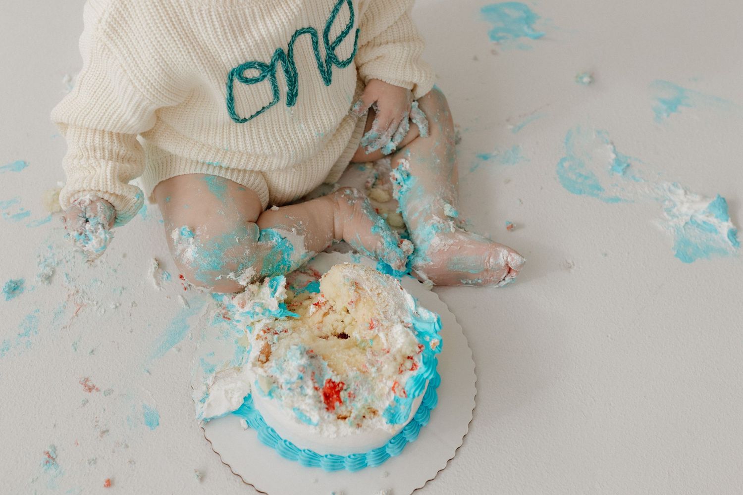Baby in white "one" sweater sitting on floor with smashed blue and white cake during first birthday cake smash photo session.