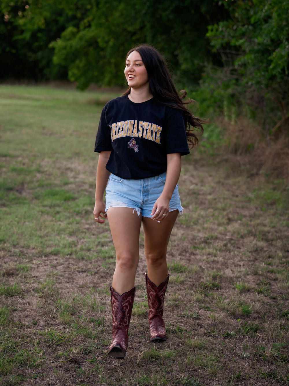 Person wearing university t-shirt and cowboy boots walking in grassy field.