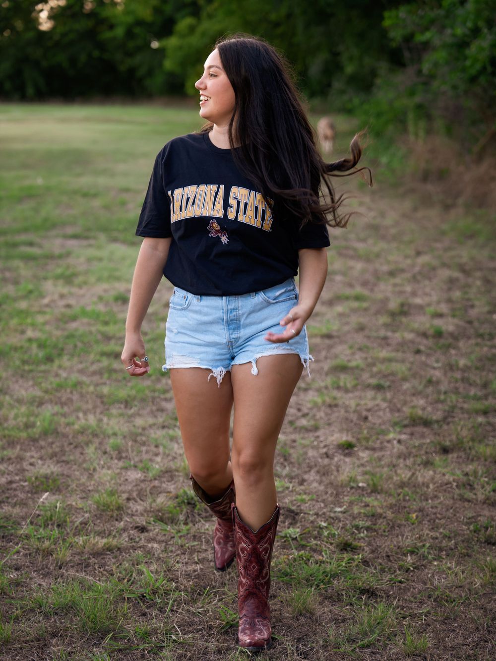 Walking through a grassy field in an Arizona State shirt, jean shorts and western boots on a sunny day.