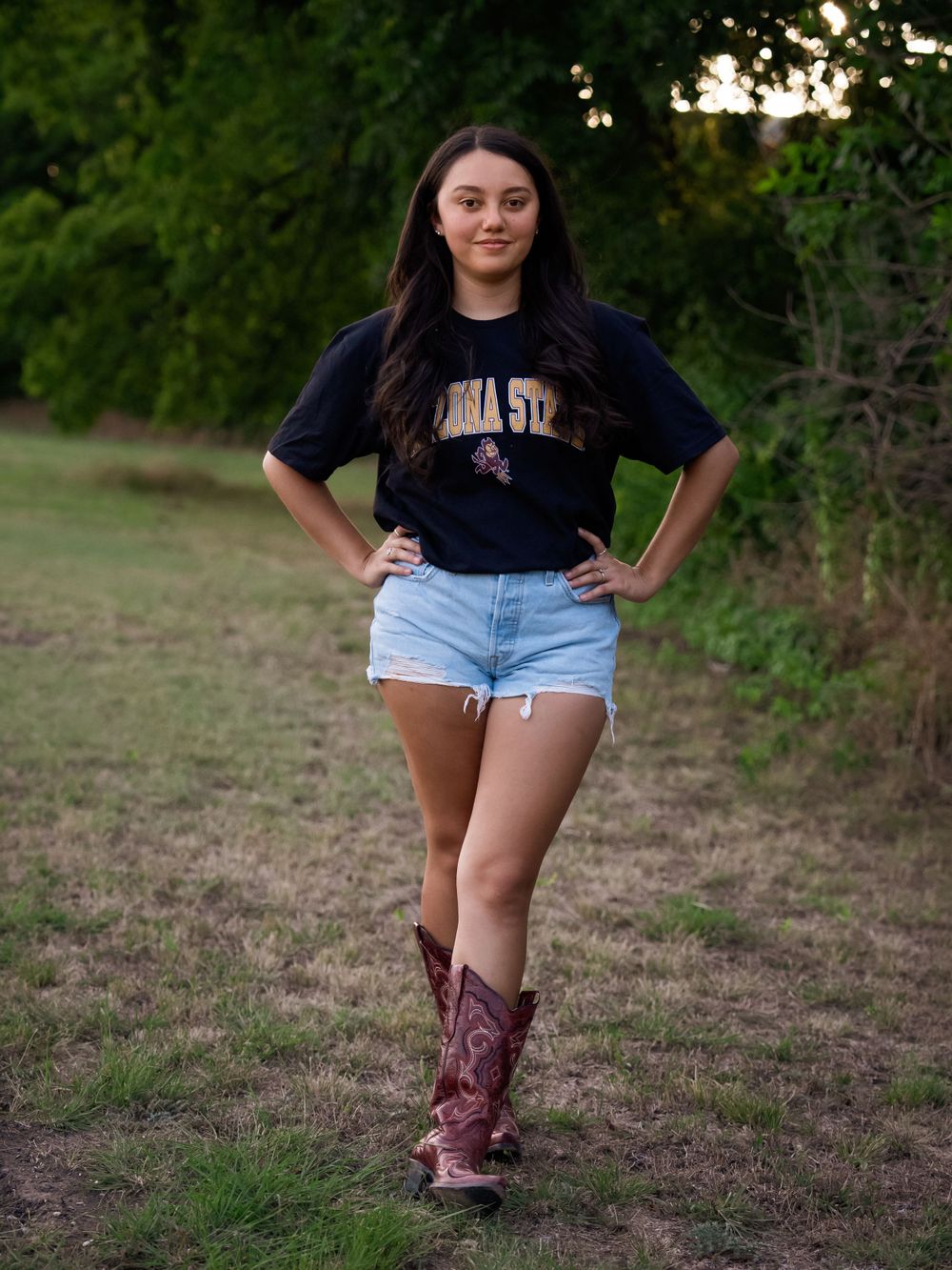 Casual outdoor portrait of someone in a university shirt and cowboy boots posing with hands on hips.