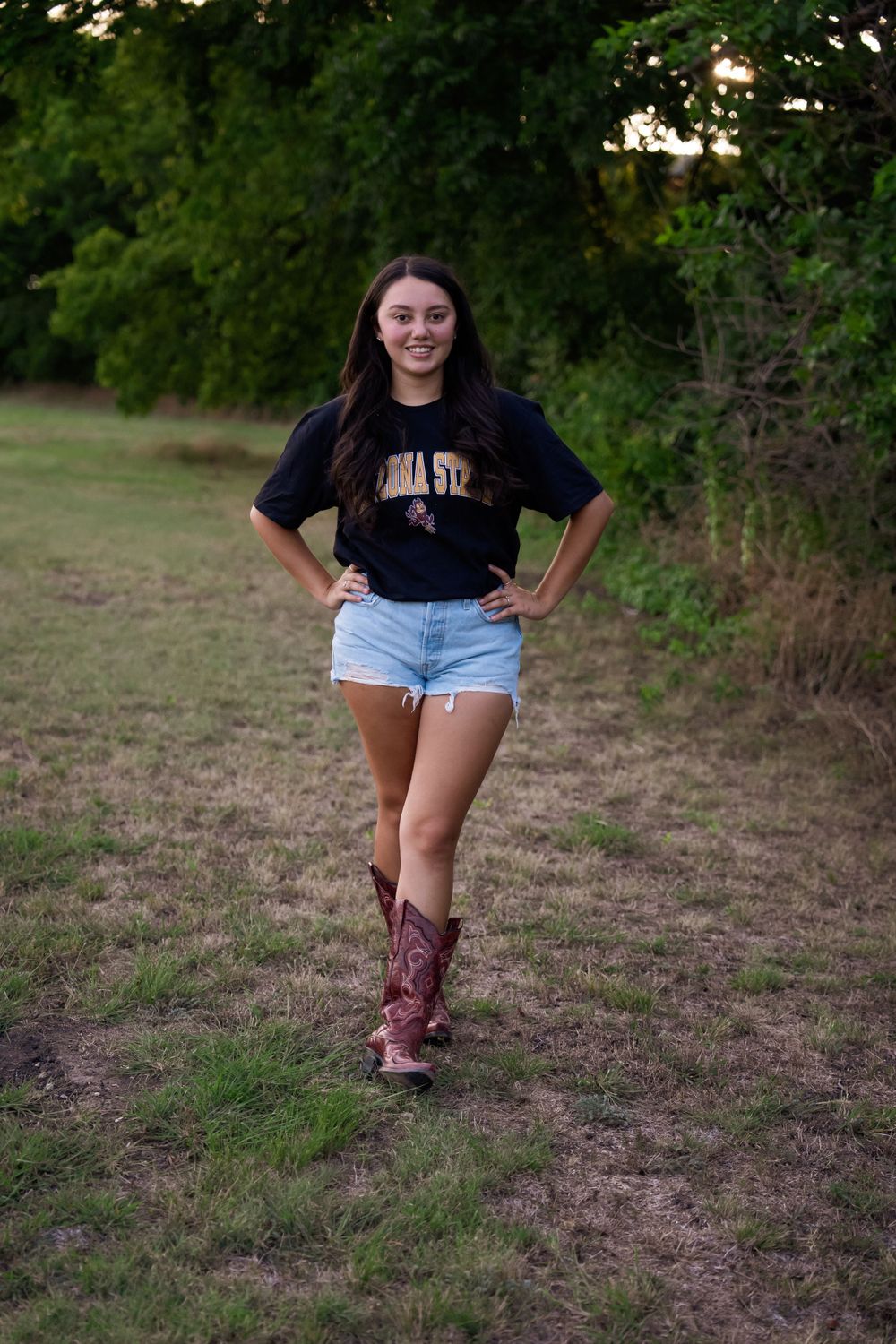 Standing outdoors wearing a collegiate t-shirt, denim shorts and embroidered cowboy boots at sunset.