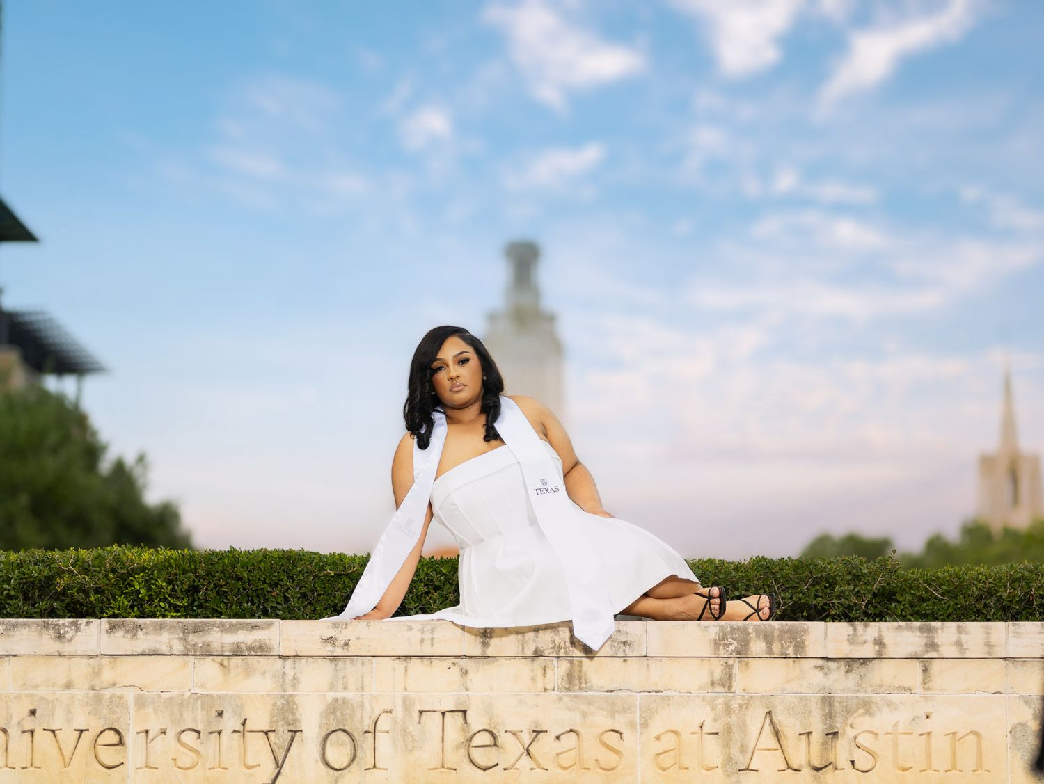 Graduate in a grey cap and gown poses outdoors on a grassy campus lawn.
