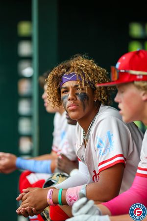 Baseball player in white uniform sitting in dugout wearing eye black and colorful accessories.