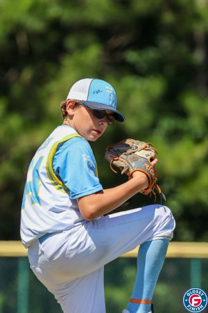 Youth baseball pitcher in light blue uniform shows perfect leg lift during windup motion on the mound.