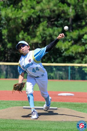 Baseball player in light blue uniform follows through on pitching motion on baseball field with trees in background.