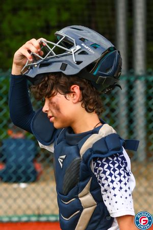 Baseball catcher in navy uniform adjusts protective gear during game break.