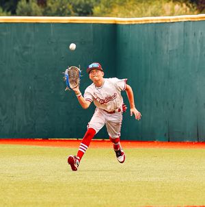 Baseball outfielder in white uniform runs to catch fly ball against green wall backdrop.