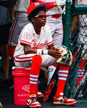 Baseball player in white and red uniform sitting in dugout with glove and equipment.