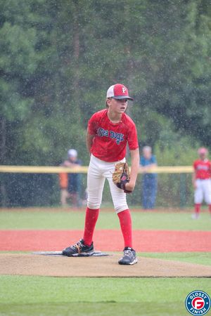 Young pitcher in red uniform demonstrates proper follow-through technique on baseball mound.