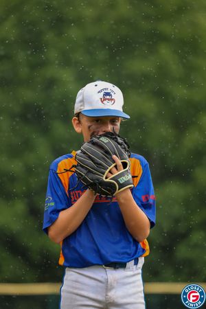 Youth baseball player in blue and orange uniform peers in for signals from behind baseball glove.