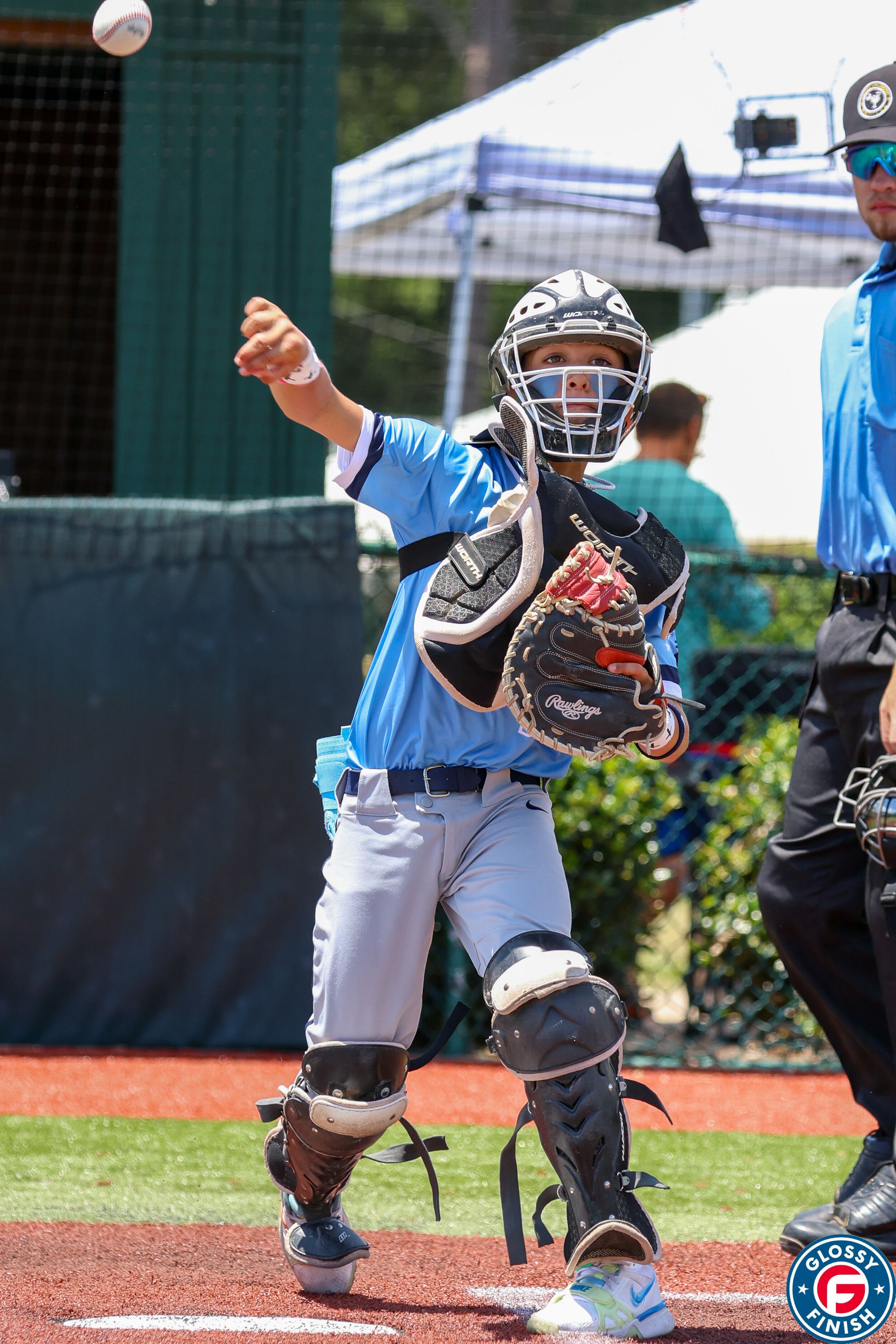 A baseball catcher in blue uniform and protective gear prepares to catch a ball during a youth baseball game.