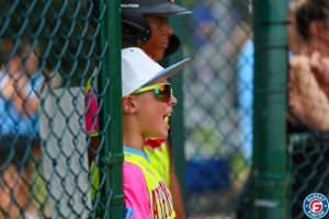 Young baseball player watching game intently from dugout wearing neon yellow jersey.
