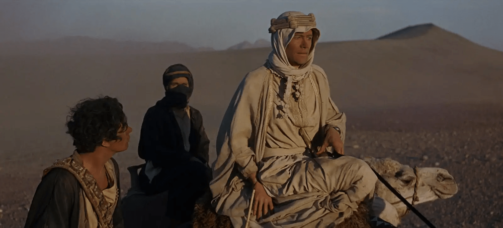 Desert travelers on camels silhouetted against vast sand dunes.