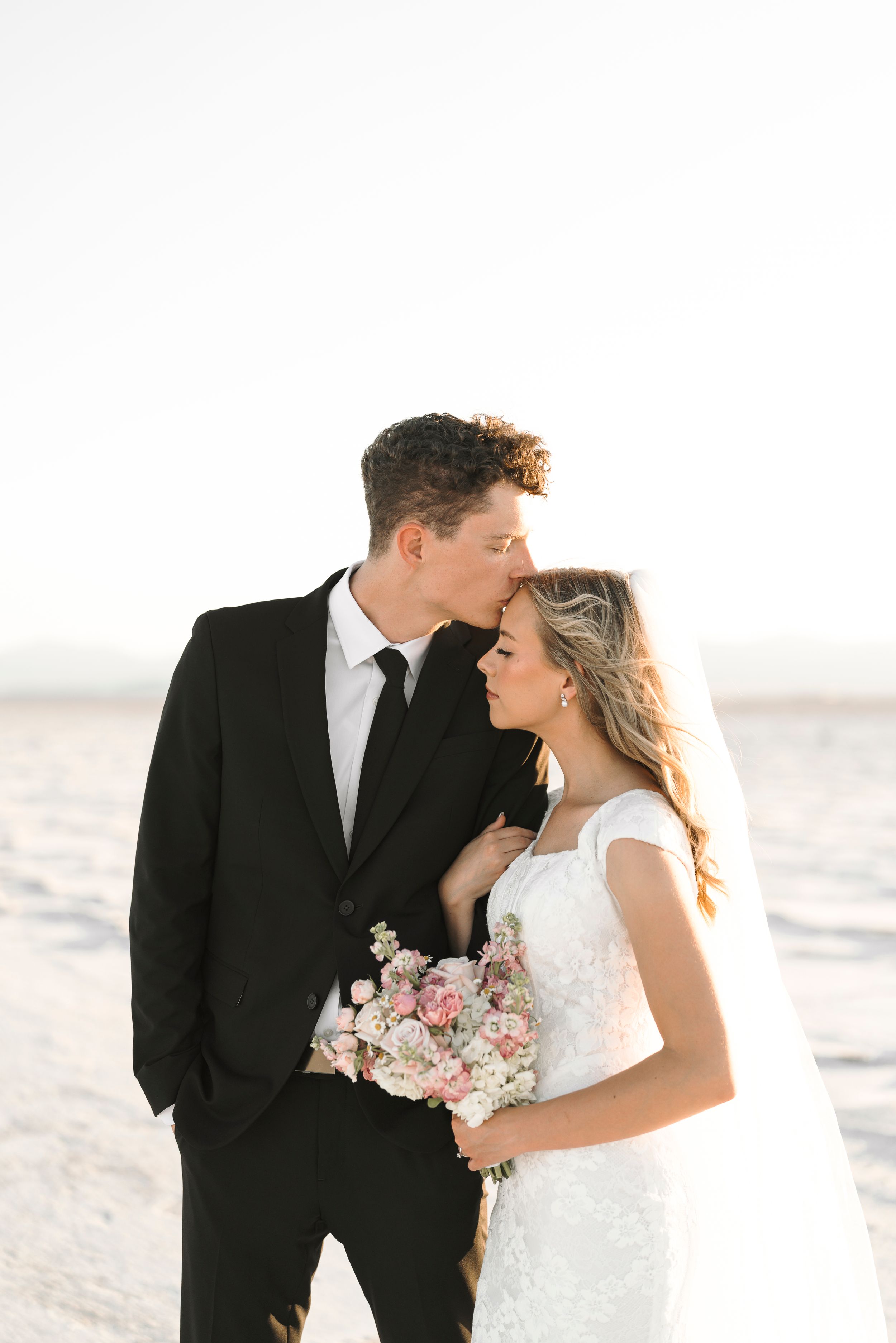 A romantic wedding portrait of a couple on a beach at sunset with the bride holding a pink and white bouquet.