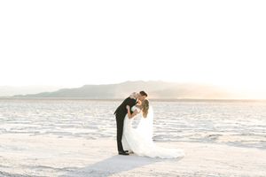 A couple in elegant wedding attire embraces against a scenic ocean backdrop at sunset.