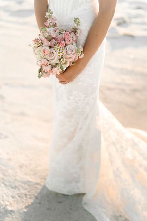 A bride in a white lace wedding dress holds a soft pink and blush rose bouquet on a sunny beach.