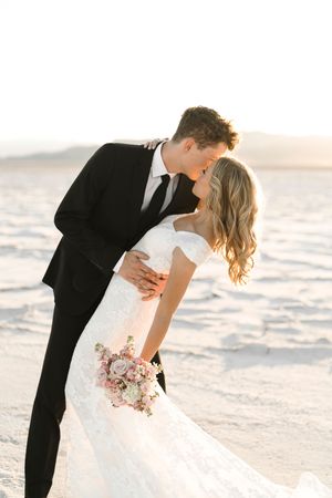 A romantic wedding couple embraces on a beach at sunset with the bride holding a pink bouquet.