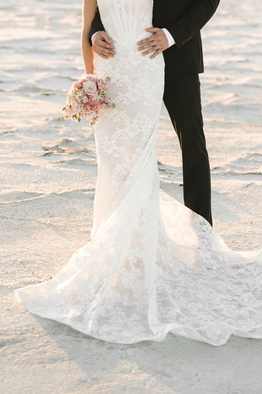 A close-up of a white lace wedding dress hemline dragging in beach sand next to black formal shoes.