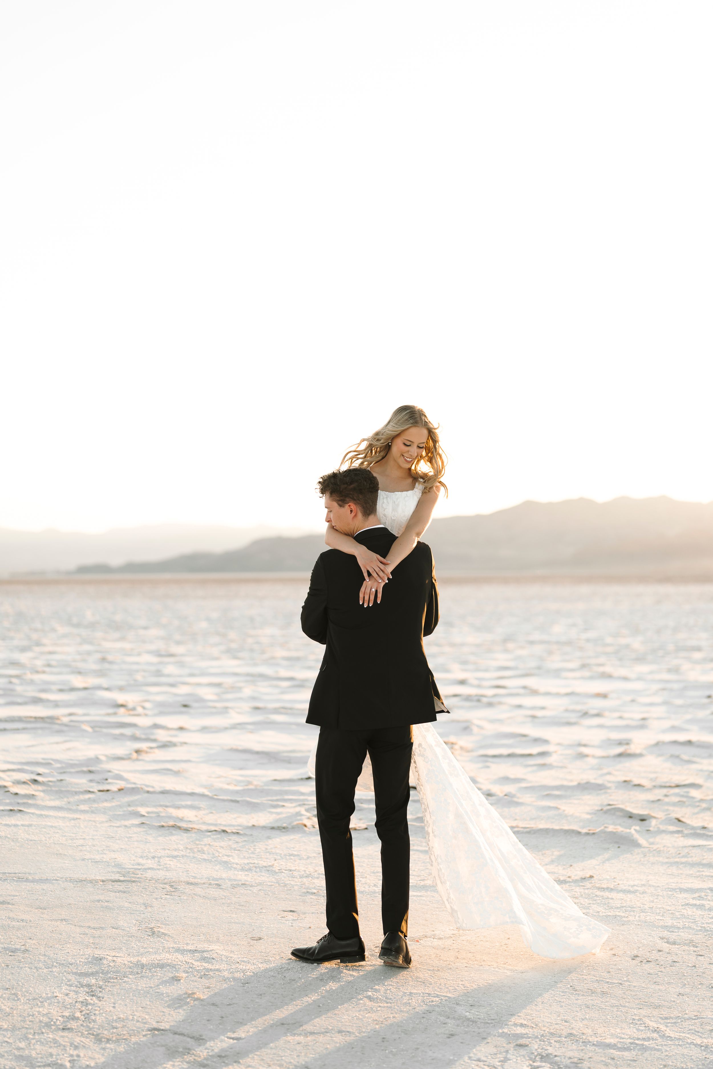 Romantic couple in black clothing embraces on white salt flats at sunset with mountains in background.