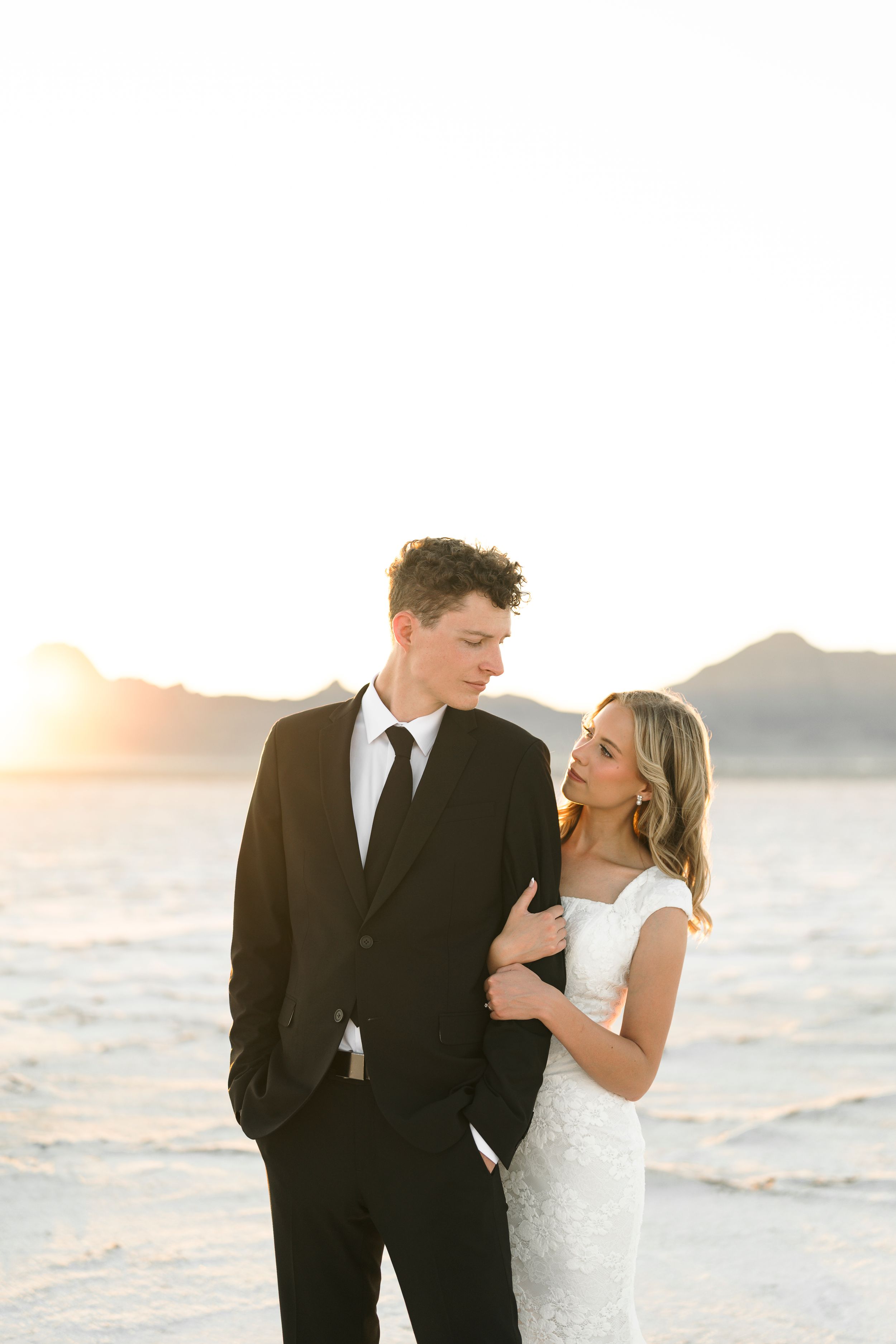 Couple in formal wedding attire embracing on a beach at sunset with mountains in the background.