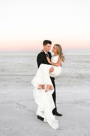 A romantic wedding couple embraces on a white sandy beach at sunset in elegant formal attire.